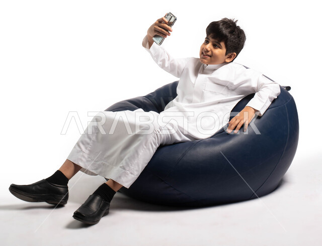 اجراء مكالمة فيديو والتواصل عن بعد , بورتريه لطفل عربي خليجي سعودي يجلس على كرسي مريح و يحمل بيده هاتف محمول , إيماءات و تعابير وجه تدل على السعادة ، أخذ صورة سيلفي تذكارية ، خلفية بيضاء