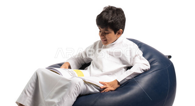 ايماءات تدل على الاستمتاع بالمطالعة, قضاء الوقت بالقراءة والتعلم والتثقف ، بورتريه لولد عربي سعودي خليجي يرتدي الزي السعودي يجلس على كرسي مريح ويحمل بيديه كتاب  ، الاستمتاع بقراءة الكتب , ثقافة التعليم الذاتي, خلفية بيضاء