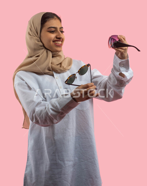 الاستمتاع في اختيار نظاره جديده مناسبة، بورتريه لإمرأه عربية سعودية خليجية بالزي الكاجوال تمسك بيديها نظارات شمسية، إيماءات وجه تدل على السعاده والفرح ،  خلفية وردية، صورة بالطول الكامل، اقتناء نظارات حماية من الشمس