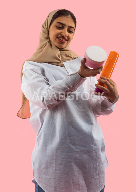  الإعلان والتسويق لمنتجات صحية، الشراء الالكتروني الاونلاين، خلفية وردية،  بورتريه لشابة  عربية خليجية سعودية، تحمل بيديها كريم الترطيب وشامبو الشعر ، مفهوم الاهتمام بالجسد، النظافة والأناقة والجمال، ايماءات الإعجاب والسعادة، التسوق من متاجر ومحلات العناي