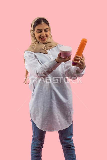 Advertising and marketing for healthy products, online online purchase, pink background, portrait of a young Saudi Arabian Gulf woman, holding moisturizing cream and hair shampoo in her hands, the concept of caring for the body, hygiene, elegance and beau