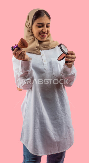  مفهوم الانوثه والاهتمام بالمظهر الخارجي, أدوات تجميل نسائيه عصريه، التسويق لمنتجات التجميل, بورتريه لفتاه عربيه سعودية خليجيه تحمل بيدها مستحضرات توريد الخدود, مكياج وميكاب تتوريال, صالون نسائي راقي, خلفيه ورديه. 