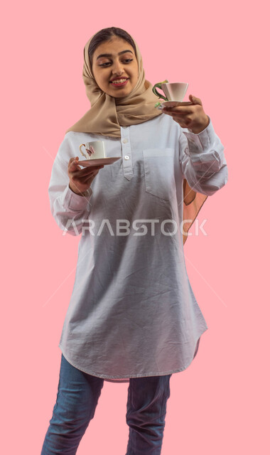  أكواب شاي و قهوة، ايماءات تدل على الحيره في اختيار الادوات المنزليه، بورتريه لفتاة عربية خليجية سعودية تحمل بيدها اواني خزفية زجاجية مزخرفة، الاستمتاع في شراء ادوات المطبخ، التسوق في محلات ومتاجر الأواني المنزليه، خلفية وردية.