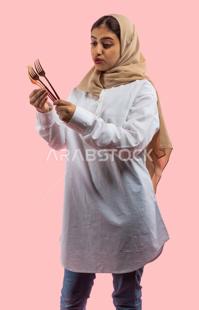  ايماءات تدل على الحيره في اختيار الادوات المنزليه،  متعة الشراء والتسوق في المحلات التجاريه, بوتريه لفتاه عربيه سعوديه خلجيه ترتدي الزي الكاجوال تمسك بيدها أدوات تناول الطعام,  معدات وتجهيزات طاولة المائده , خلفيه ورديه. 