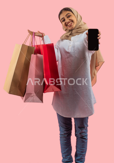 الشراء الإلكتروني الآمن، ايماءات وجه تدل على الإستمتاع بالتسوق، بورتريه لإمرأة عربية سعودية خليجية مبتسمة ترتدي الزي الكاجوال تحمل بيدها حقائب التسوق وهاتف محمول بشاشة سوداء فارغة، عروض وتخفيضات الجمعة البيضاء ، التسوق في المول التجاري ، خلفية وردية. 