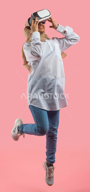 Gestures indicating happiness and integration with the imaginary world, a portrait of a Saudi Arabian Gulf girl wearing casual clothes and 3D virtual reality glasses and jumping excitedly, watching movies via technology, full-length body image, pink backg