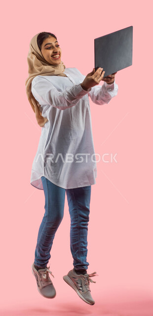 إستخدام اجهزة تقنية حديثة،  بورتريه لامرأة عربية سعودية خليجية ترتدي الزي الكاجوال تستخدم جهاز التابلت ، التسوق الإلكتروني الآمن، تصفح الإنترنت وإنجاز المهام عن بعد ، الشراء والبيع عبر الاونلاين، صورة الجسم بالطول الكامل، خلفية وردية. 