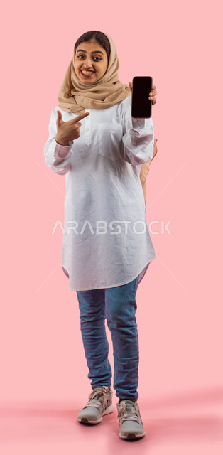 Smart phone applications, a portrait of a smiling Saudi Arabian Gulf woman holding and pointing with her hand at a mobile phone with a blank black screen, online shopping through smart phone applications and programs, marketing advertisements, full-length