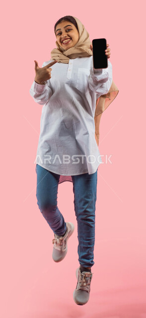 Body language and facial expressions indicating joy and pleasure in owning a new mobile phone, smartphone applications, a portrait of a smiling Saudi Arabian Gulf woman holding and pointing with her hand at a mobile phone with a blank black screen, market