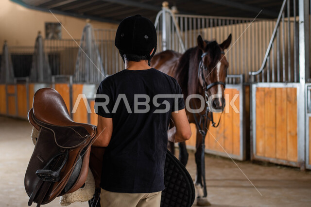 الاستعداد لركوب الخيل , الرفق والاحسان الى الحيوانات ,  ترويض وتدريب وتجهيز الخيول في الاسطبل  ، صورة من الخلف لمدرب أحصنة سعودي خليجي يرتدي زي الفروسية يحمل بيده سرج  الفرس ويقوم بتحضيره