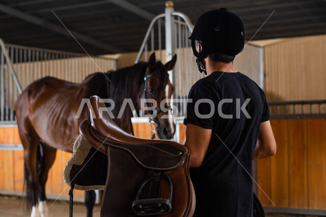 ترويض وتجهيز الخيول في الاسطبل , مهارات وأنشطة ترفيهية في مزرعة الفروسية , الرفق والإحسان إلى الحيوانات ، صورة من الخلف لرجل سعودي خليجي يرتدي زي الفروسية يحمل بيده سرج الحصان ويقوم بتجهيزه, الاستعداد لركوب الفرس