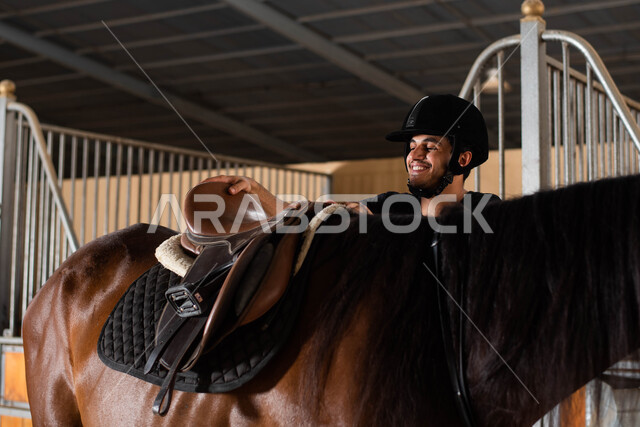 ايماءات تدل على السعادة و الاستمتاع مع الحيوانات , مفهوم الرفق والإحسان إلى الحيوان، ترويض وتدريب الخيول في اسطبل الأحصنة ، جواد عربي بني أصيل في مزرعة تربية الجياد, رجل سعودي خليجي عربي يرتدي زي الفروسية و يقوم بتركيب السرج على ظهر  الفرس