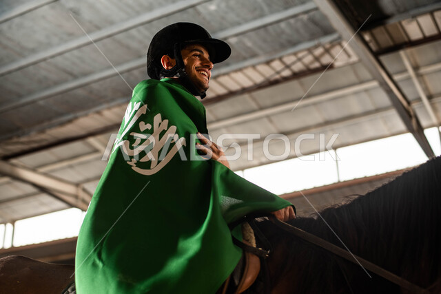 جواد عربي بني أصيل , رعاية وتدريب الأحصنة , أنشطة ترفيهية في اسطبل الخيول , هواية ركوب الخيل, صورة جانبية لشاب عربي سعودي خليجي يرتدي علم السعودية على ظهره ويمتطي حصانه , الاحتفال باليوم الوطني السعودي 2023 , ذكرى يوم التأسيس, يوم العلم السعودي