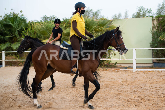 أنشطة وفعاليات ترفيهية بين الأصدقاء برفقة الأحصنة، شاب و شابة عربيان خليجيان سعوديان يمتطيان خيولهما العربية الأصيلة، مزرعة سعودية لتدريب وترويض الخيول في الاسطبل، ممارسة هواية ركوب الخيل، الإلتزام بالاحترازات الوقائية بإرتداء الكمامة،  الوقاية من الأمراض