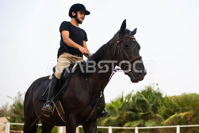 صورة جانبية لحصان أصيل أسود اللون يقوده شاب عربي سعودي خليجي يرتدي زي الفروسية، رياضات و فعاليات ترفيهية ضمن مزرعة سعودية لتدريب وترويض الخيول، ممارسة هواية ركوب الخيل في الإسطبل،  قضاء أوقات ممتعة في التدريب على ركوب الجواد. 