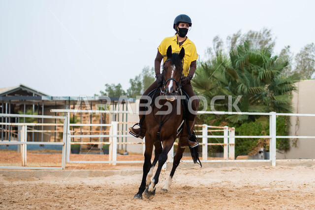 قضاء أوقات ممتعة برفقة الحصان ، مزرعة تدريب وترويض الخيول، اسطبل الأحصنة في المملكة العربية السعودية، هواية ركوب الخيل ، شابة عربية سعودية خليجية ترتدي زي الفروسية وتمتطي الجواد العربي البني، الإلتزام بالاحترازات الوقائية بإرتداء الكمامة. 