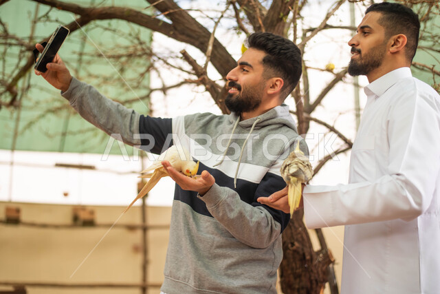 Documenting moments and taking memorial photos using a mobile phone, two Saudi Arabian Gulf men spending fun times in the zoo, recreational activities with birds, the Riyadh season safari event.
