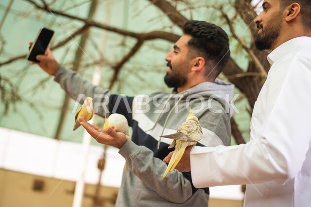 توثيق اللحظات والتقاط الصور التذكارية باستخدام الهاتف المحمول، رجلان عربيان خليجيان سعوديان يمضيان اوقات ممتعة في حديقة الحيوانات ، أنشطة ترفيهية مع الطيور ، فعالية سفاري موسم الرياض.