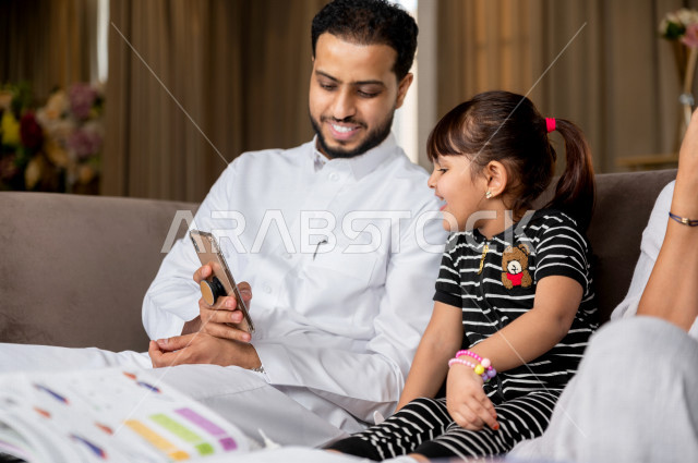 تلميذة سعودية خليجية تدرس في المنزل مع والدها و والدتها ، يساعدانها على تأدية واجباتها المدرسية ، استخدام الهاتف المحمول لمتابعة التعليم ، مفهوم التعليم عبر الانترنت ، التعليم المنزلي اثناء الحجر الصحي و تفشي فيروس كورونا ، العزلة الذاتية .