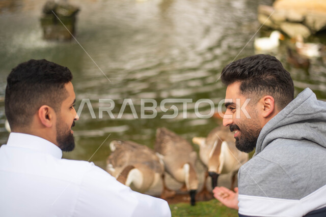 قضاء اوقات ممتعة داخل حديقة الحيوانات في المملكة العربية السعودية، فعالية سفاري موسم الرياض، رجلان عربيان خليجيان سعوديان يطعمان طائر البط عند بركة المياه في محمية طبيعية.