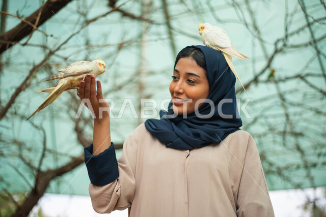 خلفية الطبيعة، إيماءات وجه تدل على السعادة والاستمتاع باللهو مع الطيور، أماكن سياحية وفعاليات ترفيهية بالسعودية، صورة مقربة لامرأة عربية خليجية سعودية محجبة في حديقة الحيوانات في المملكة العربية السعودية، محمية طبيعية.