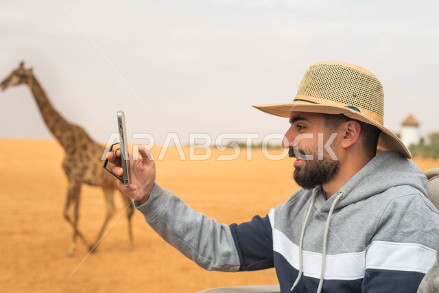توثيق اللحظات و التقاط صور تذكارية بإستخدام الهاتف, رجل عربي خليجي سعودي يقوم بجولة سياحية في حديقة الحيوانات في المملكة العربية السعودية, قضاء أوقات ممتعة, ايماءات وجه تدل على السعادة و الاستمتاع, أنشطة و فعاليات ترفيهية في موسم سفاري الرياض.
