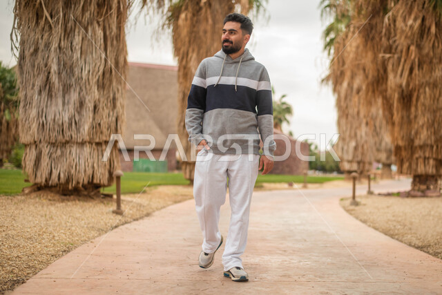 شاب عربي خليجي سعودي يتجول في إحدى الحدائق العامة، قضاء أوقات ممتعة، التجول و المشي في الهواء الطلق في حدائق ومنتزهات المملكة العربية السعودية, صورة الجسم بالطول الكامل.