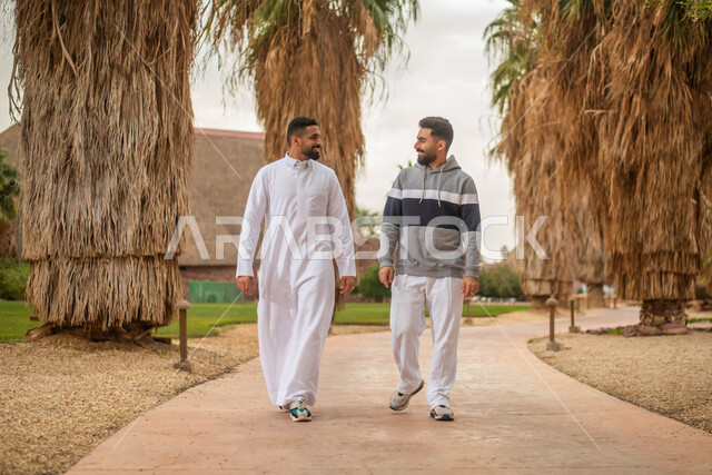 شابان عربيان خليجيان سعوديان يتجولان في احدى الحدائق العامة، قضاء أوقات ممتعة برفقة الأصدقاء، ايماءات وجه تدل على السعادة و السرور، نقاشات وحوارات و تبادل الأحاديث، نزهه في الهواء الطلق في حدائق ومنتزهات المملكة العربية السعودية, صورة الجسم بالطول الكامل.