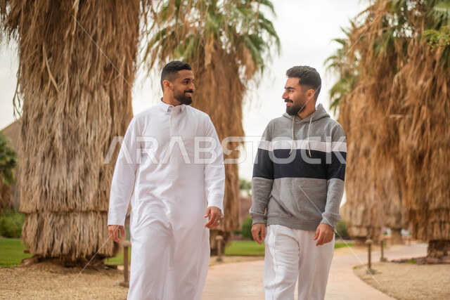 Two young Saudi Arabian Gulf men wandering in a public park, spending fun times with friends, facial gestures indicating happiness and pleasure, discussions, dialogues and exchanging conversations, a picnic in the open air in the gardens and parks of the