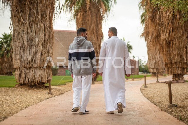 قضاء أوقات ممتعة برفقة الأصدقاء، التنزه في الهواء الطلق في حدائق ومنتزهات المملكة العربية السعودية, صوره من الخلف لشابان عربيان خليجيان سعوديان يتجولان في احدى الحدائق العامة،  نقاشات وحوارات و تبادل الأحاديث, صورة الجسم بالطول الكامل.
