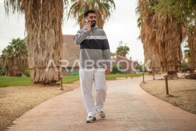 رجل عربي خليجي سعودي يقوم بإجراء اتصال هاتفي و التجول في إحدى الحدائق العامة في المملكة العربية السعودية، استخدام الهاتف المحمول، قضاء أوقات ممتعة في الطبيعة الخضراء، نزهه في الهواء الطلق, صورة الجسم بالطول الكامل.