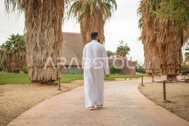 التجول و المشي في الهواء الطلق في حدائق ومنتزهات المملكة العربية السعودية, صورة من الخلف لشاب عربي خليجي سعودي يتجول في إحدى الحدائق العامة، قضاء أوقات ممتعة، صورة الجسم بالطول الكامل.