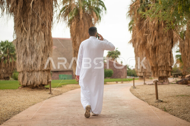 قضاء أوقات ممتعة في الطبيعة الخضراء، استخدام الهاتف المحمول، صورة من الخلف رجل عربي خليجي سعودي يقوم بإجراء اتصال هاتفي و التجول في إحدى الحدائق العامة في المملكة العربية السعودية، نزهه في الهواء الطلق, صورة الجسم بالطول الكامل.