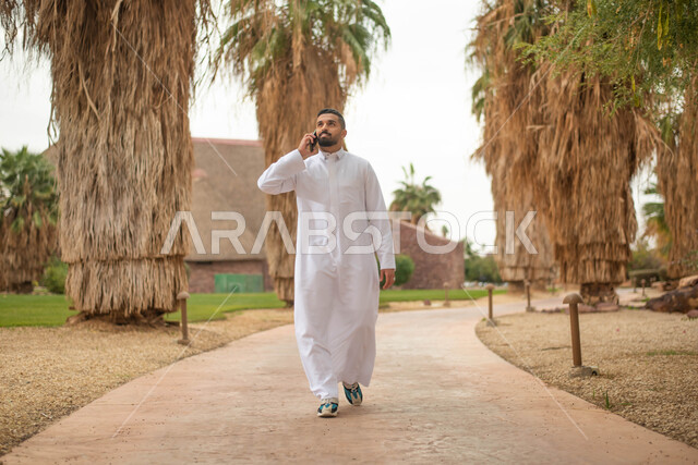 استخدام الهاتف المحمول في الرد على الاتصالات الهاتفية، رجل عربي خليجي سعودي يقوم بإجراء اتصال هاتفي و التجول في إحدى الحدائق العامة في المملكة العربية السعودية،  قضاء أوقات ممتعة في الطبيعة الخضراء، نزهه في الهواء الطلق, صورة الجسم بالطول الكامل.