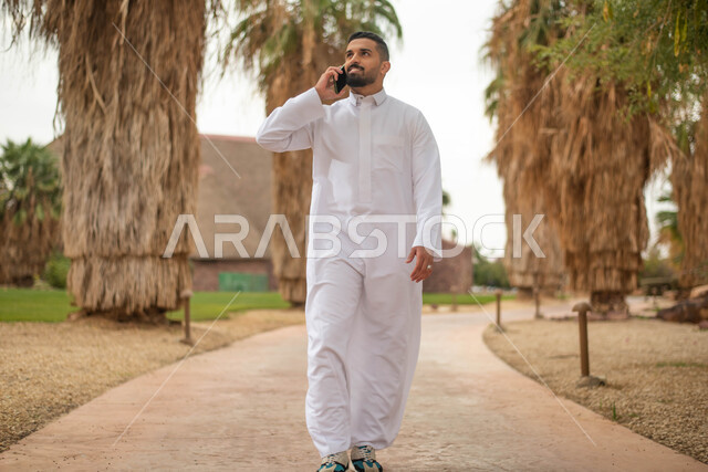 رجل عربي خليجي سعودي يقوم بإجراء اتصال هاتفي و التجول في إحدى الحدائق العامة في المملكة العربية السعودية، استخدام الهاتف المحمول، قضاء أوقات ممتعة في الطبيعة الخضراء، نزهه في الهواء الطلق, صورة الجسم بالطول الكامل.