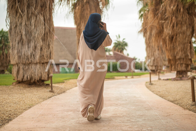 نزهه في الهواء الطلق، جولة في الطبيعة الخضراء, إجراء مكالمه هاتفيه عبر الهاتف المحمول، صورة من الخلف لإمرأة عربية خليجية سعودية تقضي أوقات ممتعة في إحدى الحدائق في السعودية، دردشه ومحادثات,  صورة الجسم بالطول الكامل.