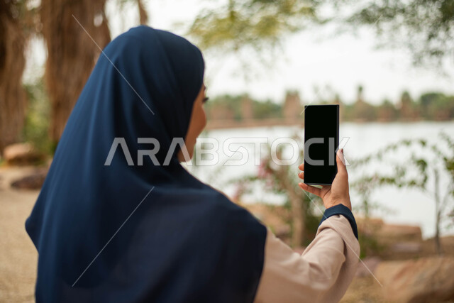 Taking souvenir photos using a mobile phone, a Saudi Arabian Gulf woman holding a phone with a blank black screen in her hand, spending enjoyable times in one of the parks of the Kingdom of Saudi Arabia, live broadcasting, hiking in the open air and green