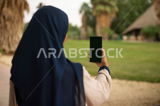 قضاء أوقات ممتعة في إحدى حدائق المملكة العربية السعودية، نزهه في الهواء الطلق و الطبيعة الخضراء, التقاط صور تذكارية باستخدام الهاتف، صورة مقربة لإمرأة عربية خليجية سعودية تحمل هاتف محمول بشاشة سوداء فارغة,  بث مباشر.
