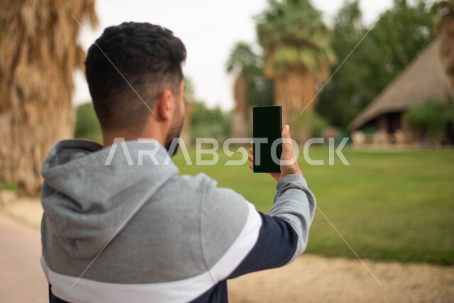 الرد على مكالمات الفيديو باستخدام الهاتف المحمول، صورة مقربة لشاب عربي خليجي سعودي يحمل بيده هاتف بشاشة سوداء فارغة يقضي أوقات ممتعة في إحدى حدائق المملكة العربية السعودية، التقاط صور تذكارية للطبيعة الخضراء ، نزهه في الهواء الطلق.