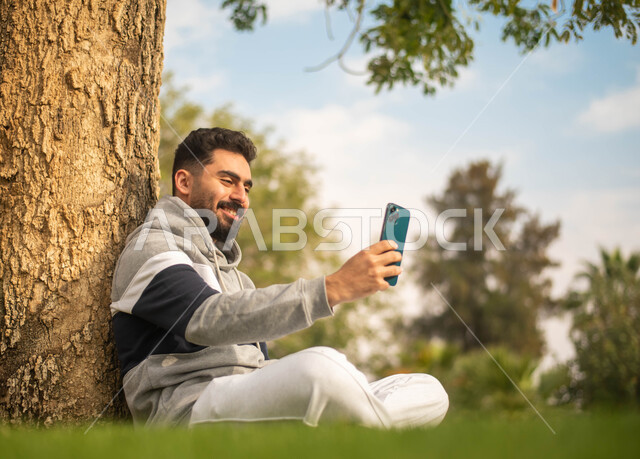 رجل عربي خليجي سعودي يجلس تحت شجرة و يقوم بإجراء الاتصالات الهاتفية, استخدام الهاتف المحمول،الحدائق العامة في المملكة العربية السعودية، إيماءات وجه تدل على السعادة، نزهه في الهواء الطلق، قضاء أوقات ممتعة في الطبيعة الخضراء.