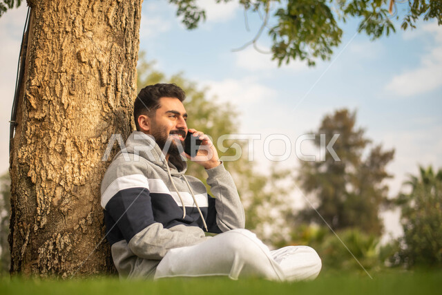 رجل عربي خليجي سعودي يجلس تحت شجرة و يقوم بإجراء الاتصالات الهاتفية, استخدام الهاتف المحمول،الحدائق العامة في المملكة العربية السعودية، إيماءات وجه تدل على السعادة، نزهه في الهواء الطلق، قضاء أوقات ممتعة في الطبيعة الخضراء.