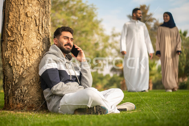 A Saudi Gulf Arab man sits under a tree and makes phone calls, using a mobile phone, public parks in the Kingdom of Saudi Arabia, facial gestures indicating happiness, strolling in the open air, spending enjoyable times in the green nature.