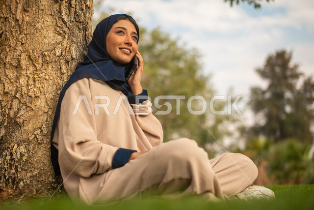 إيماءات وجه تدل على السعادة، الرد على المكالمات الهاتفية, صورة مقربة لإمرأة عربية خليجية سعودية محجبة تجلس تحت شجرة في حديقة عامة و تستخدم الهاتف المحمول، نزهه في الهواء الطلق، قضاء أوقات ممتعة في الطبيعة الخضراء.
