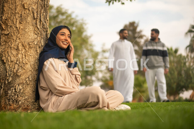 قضاء وقت ممتع في الطبيعة الخضراء وقت الصباح, إمرأة عربية خليجية سعودية محجبة تجلس تحت شجرة في حديقة عامة تجري مكالمة هاتفية عبر الجوال، إيماءات وجه تدل على السعادة, شابان عربيان سعوديان خليجيان يقومان بنزهه في الهواء الطلق, خلفية الطبيعة.