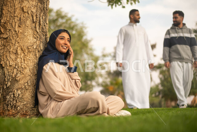 قضاء أوقات ممتعة في الطبيعة الخضراء, الرد على المكالمات الهاتفية, إمرأة عربية خليجية سعودية محجبة تجلس تحت شجرة في حديقة عامة و تستخدم الهاتف المحمول، إيماءات وجه تدل على السعادة، شابان عربيان خليجيان سعوديان يقومان بالتجول في الهواء الطلق.