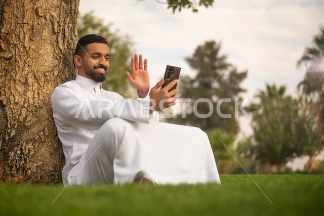 رجل عربي خليجي سعودي يجلس تحت شجرة و يقوم بإجراء الاتصالات الهاتفية, استخدام الهاتف المحمول،الحدائق العامة في المملكة العربية السعودية، إيماءات وجه تدل على السعادة، نزهه في الهواء الطلق، قضاء أوقات ممتعة في الطبيعة الخضراء.