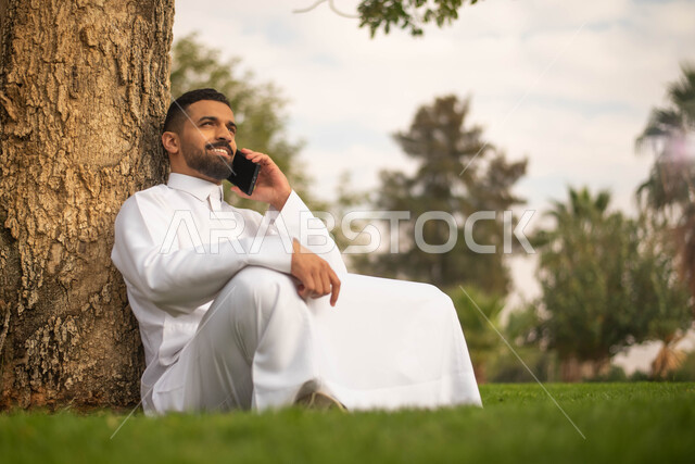 رجل عربي خليجي سعودي يجلس تحت شجرة و يقوم بإجراء الاتصالات الهاتفية, استخدام الهاتف المحمول،الحدائق العامة في المملكة العربية السعودية، إيماءات وجه تدل على السعادة، نزهه في الهواء الطلق، قضاء أوقات ممتعة في الطبيعة الخضراء.