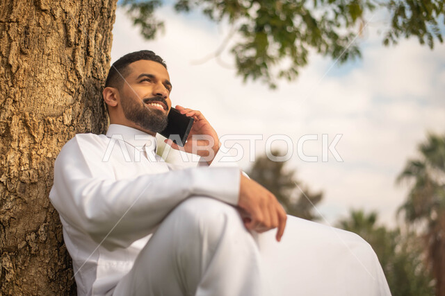 رجل عربي خليجي سعودي يجلس تحت شجرة و يقوم بإجراء الاتصالات الهاتفية, استخدام الهاتف المحمول،الحدائق العامة في المملكة العربية السعودية، إيماءات وجه تدل على السعادة، نزهه في الهواء الطلق، قضاء أوقات ممتعة في الطبيعة الخضراء.