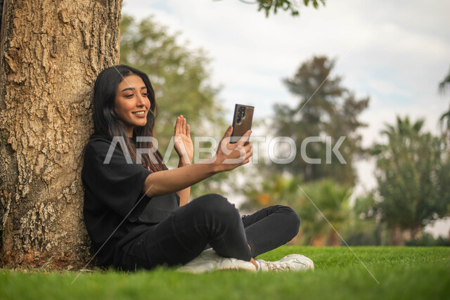 A video call, a young Saudi Arabian Gulf woman sitting on the grass and using a mobile phone, public parks in the Kingdom of Saudi Arabia, facial and hand gestures indicating happiness, a picnic in the open air, spending enjoyable times in the green natur