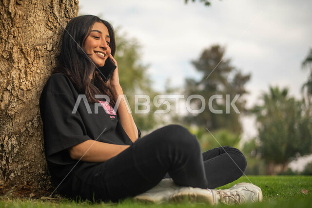 إستخدام الهاتف المحمول, صورة جانبية لشابة عربية خليجية سعودية تجلس تحت شجرة في حديقة عامة و تقوم بالرد على الاتصالات الهاتفية, إيماءات وجه تدل على السعادة، نزهه في الطبيعة الخضراء، قضاء أوقات ممتعة في الهواء الطلق.